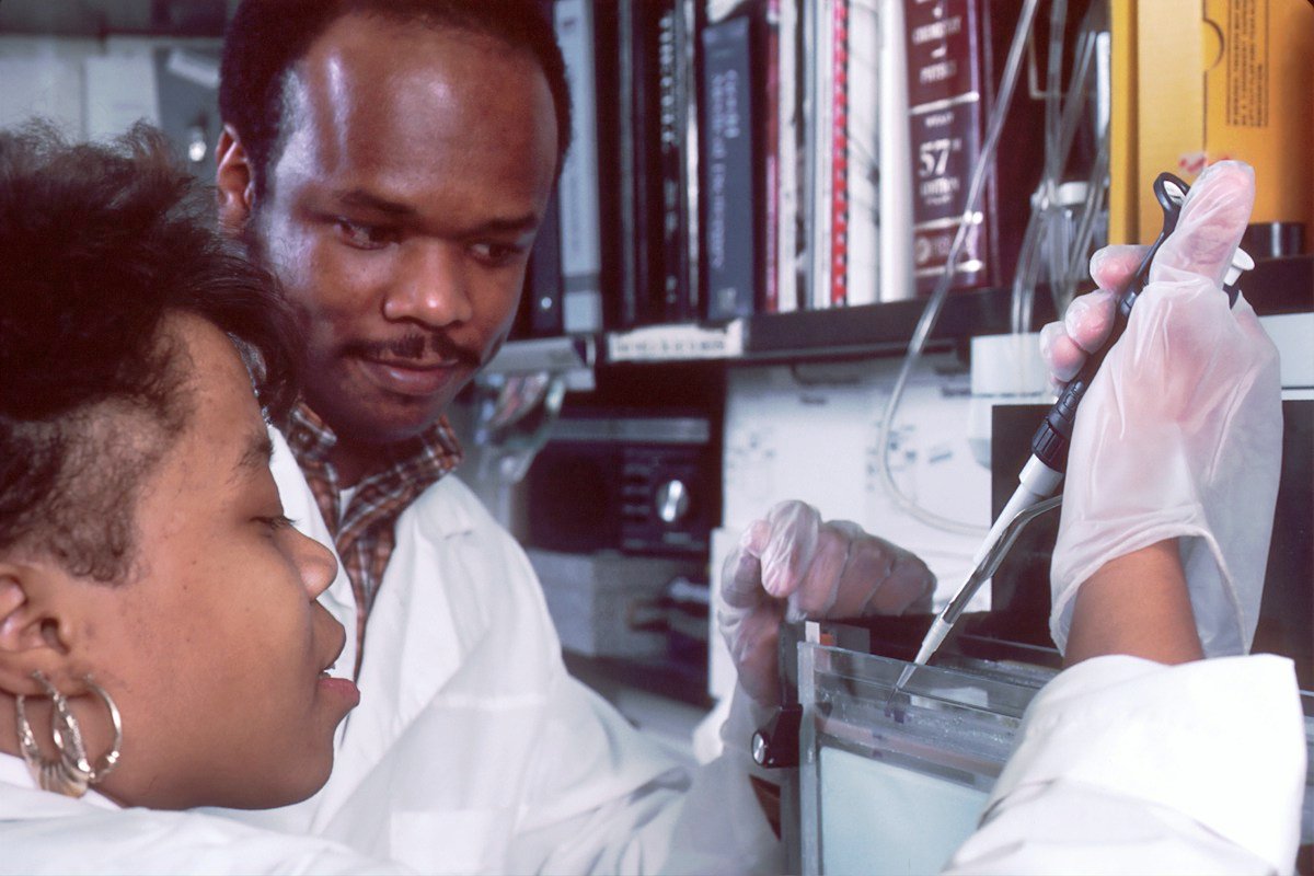 A Black scientist demonstrating a laboratory technique to a Black student; illustrative research training photo, not a scene from Flint Medical College or Xavier Ochsner