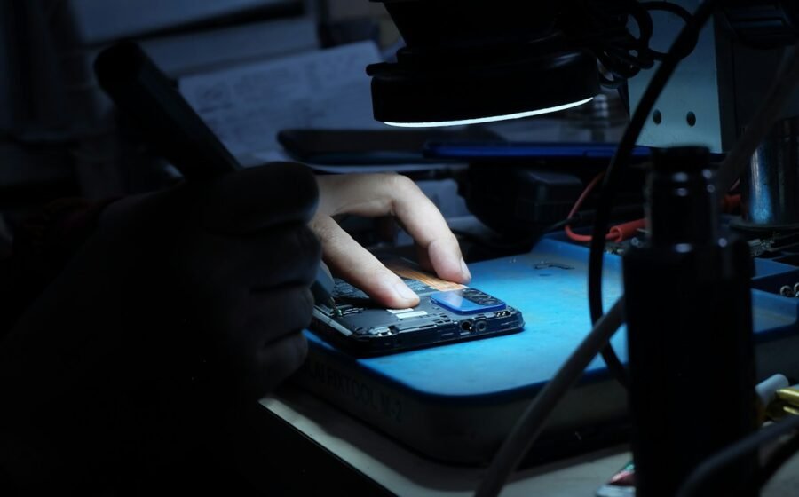 Illustrative photograph of a technician inspecting a smartphone circuit board under magnification, accompanying investigation into iPhone Duo hinge and display failures; not the iPhone Duo or an Apple/Foxconn facility
